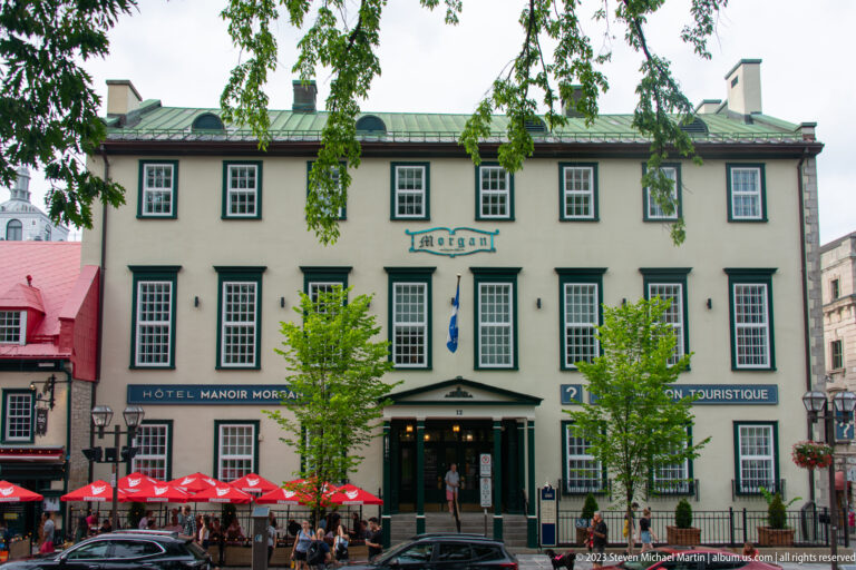 Stone façade of Hôtel Manoir Morgan with dark window trim, green roof, street trees, café umbrellas, cars, and pedestrians.