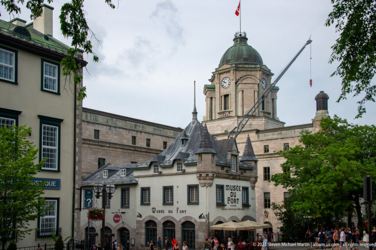 Domed clock tower behind steep-roofed historic buildings, shops, trees, and crowds on an Old Québec street.