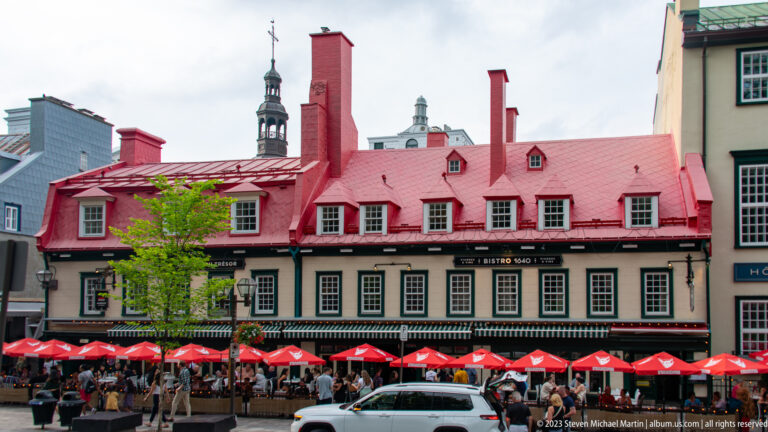 Long historic building with bright red metal roof, dormers and chimneys above a crowded restaurant terrace with red umbrellas.