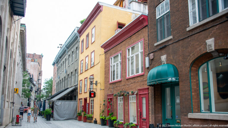 Narrow Quebec City street lined with brick and brightly painted façades, storefront awnings, plants, and pedestrians.