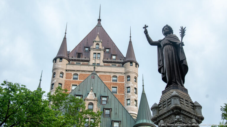 Robed bronze statue holding a cross and palm frond beside the steep roofs and turrets of Château Frontenac.