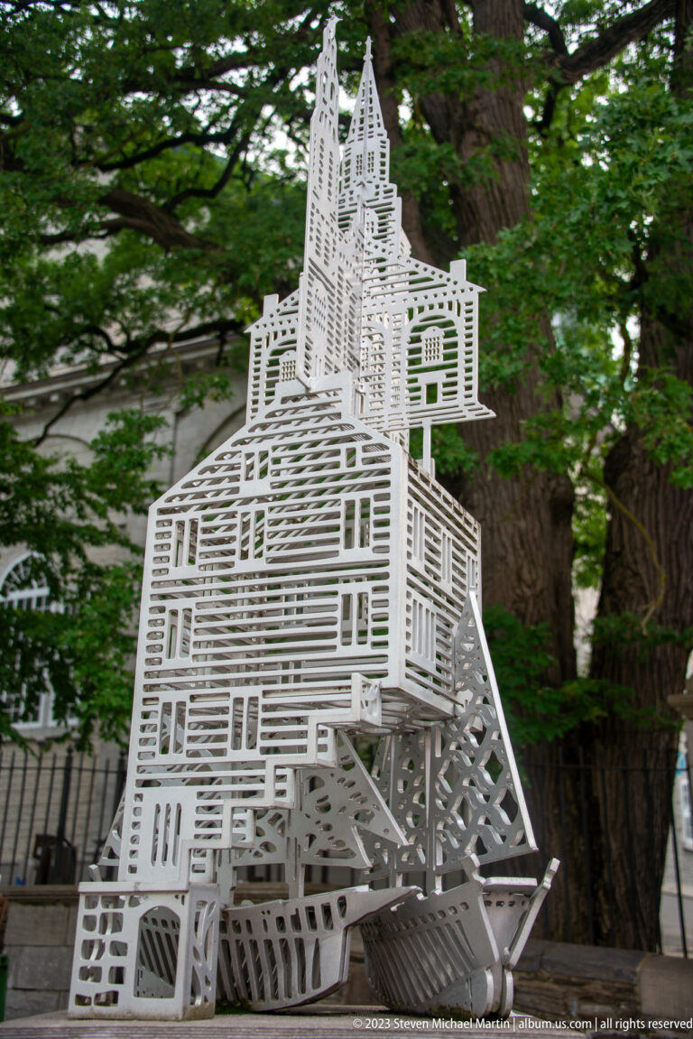 White openwork sculpture shaped like a tall historic building, formed from layered linear frames among green trees.
