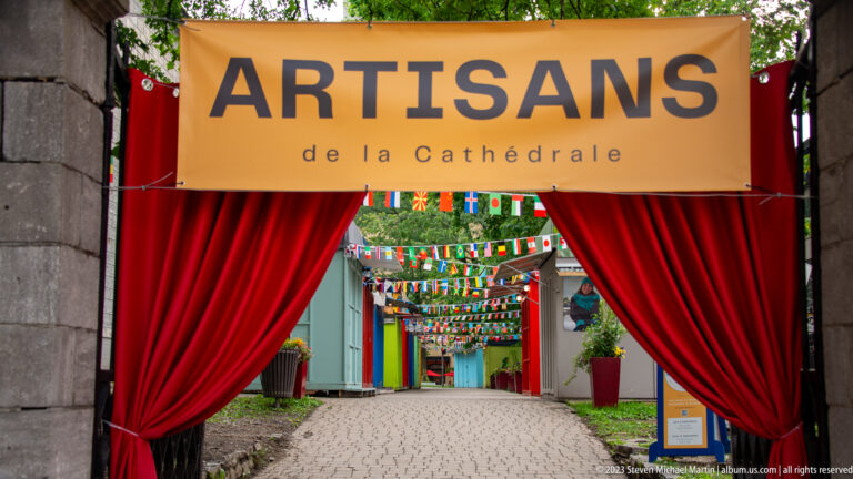 Entrance marked Artisans de la Cathédrale, framed by red curtains and opening to a cobbled lane with colorful overhead pennants.