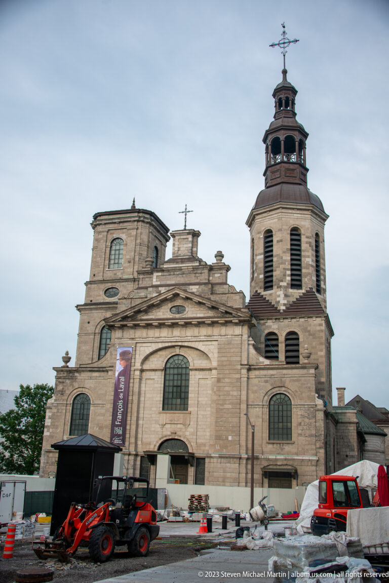 Large stone church with broad façade and tall open bell tower, with construction vehicles and barriers across the foreground.