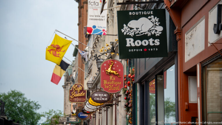 Rows of hanging shop signs, including Roots and smaller boutique signs, project over a narrow Old Québec commercial street.