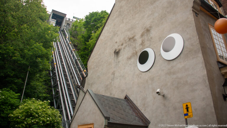 Steep inclined metal rail structure rising beside a pale wall with two circular features and dense green trees.