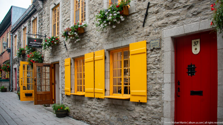 Rough stone façade with bright yellow shutters and windows, flower boxes, an open glazed door, and red door numbered 37.