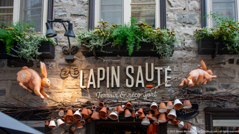 Lapin Sauté sign on a stone wall, decorated with sculpted rabbits, greenery, branches, terracotta pots, and planted window boxes.