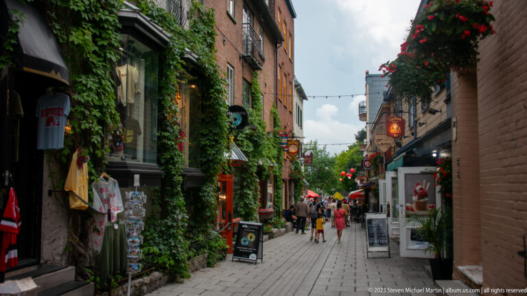 Narrow stone-paved pedestrian lane lined with shops, hanging signs, climbing greenery, flowers, display racks, and visitors.
