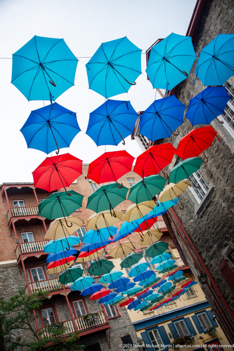 Multicolored open umbrellas suspended in rows above a narrow street between historic stone and brick buildings.