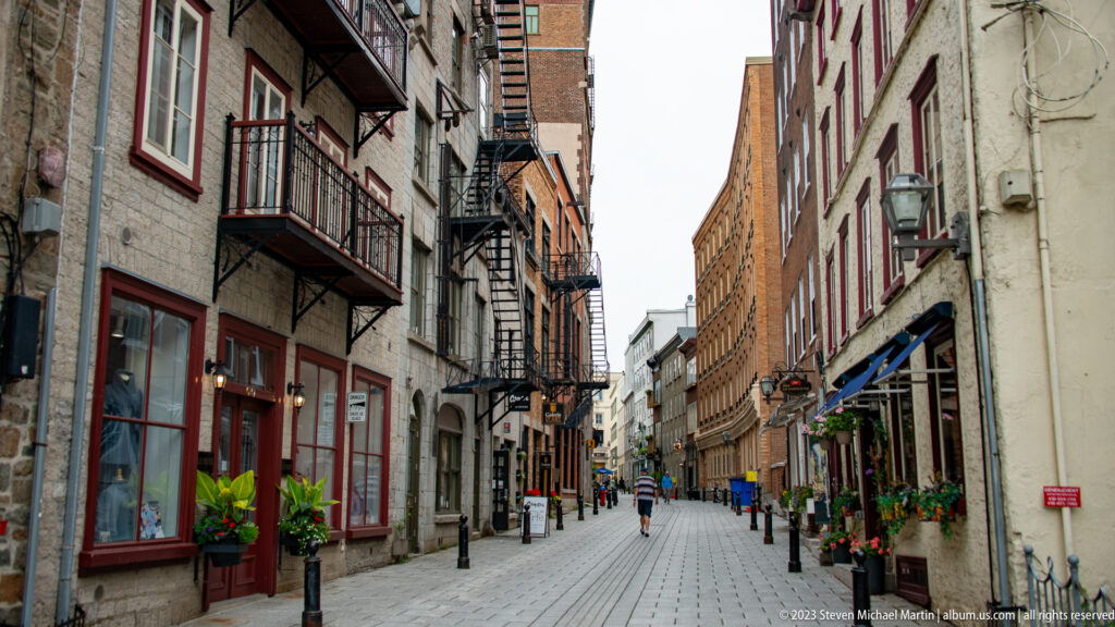Long narrow lane between stone and brick buildings with balconies, fire escapes, shops, bollards, planters, and pedestrians.