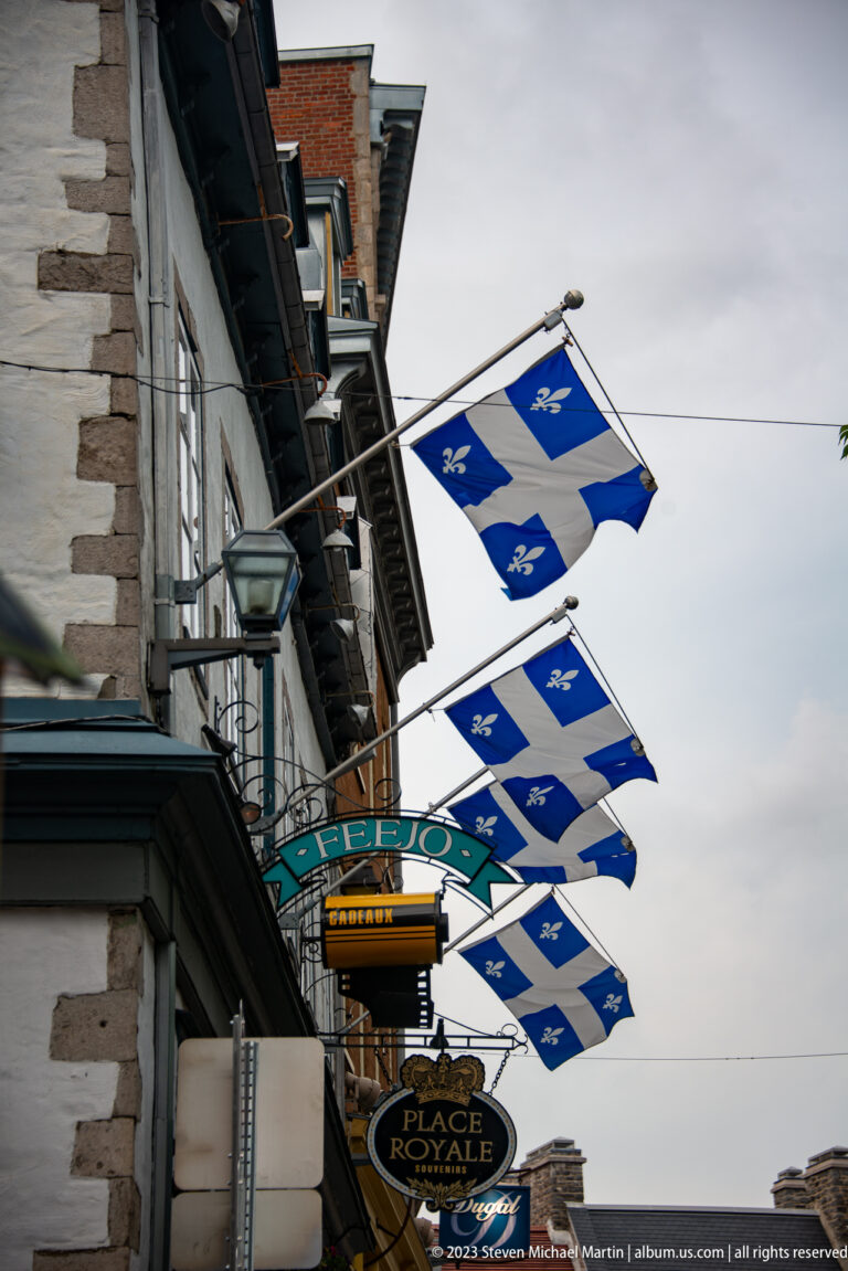 Rows of blue Québec flags hang above historic stone façades and a projecting sign reading Place Royale.