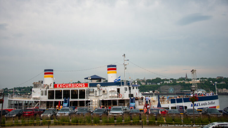 AML Louis Jolliet excursion vessel at the waterfront with twin funnels, EXCURSIONS sign, parked cars, and distant riverbank.