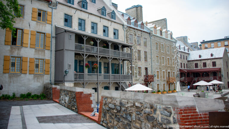 Enclosed paved courtyard surrounded by historic stone buildings with wooden galleries, dormers, shutters, retaining walls, and café umbrellas.