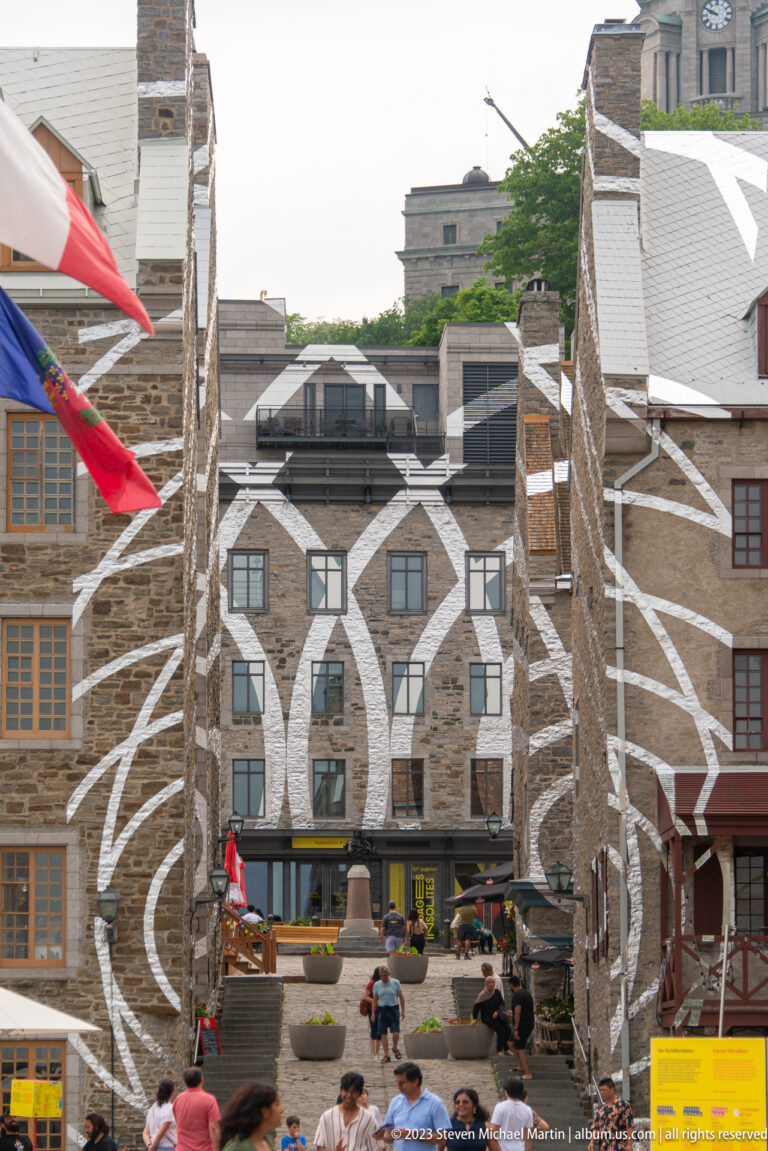 Large black-and-white linear mural spans several stone façades and windows above people gathered in an open courtyard.