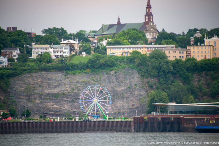 Across the river, a Ferris wheel sits below a steep bluff topped by a large church and other buildings.