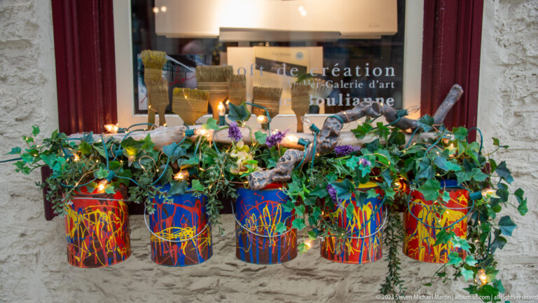 Five brightly painted cylindrical planters filled with greenery and decorations beneath a red-framed art-gallery window on a stone wall.