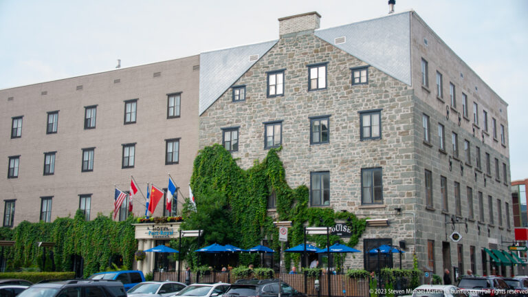 Large stone hotel with gabled historic façade, ivy, flags, blue café umbrellas, parked cars, and a taller rear wing.