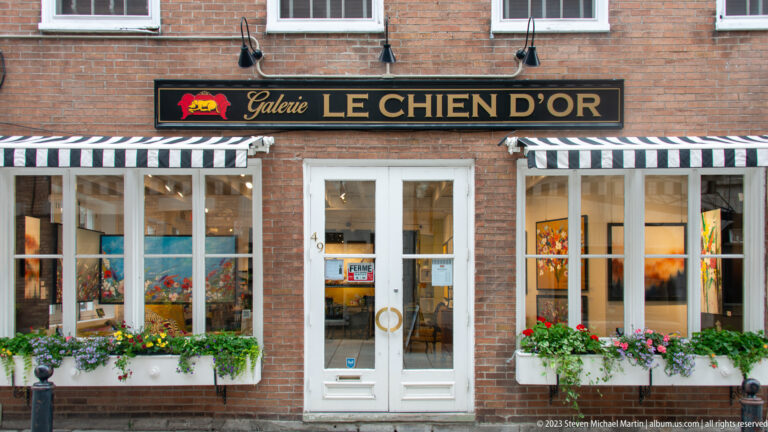 Brick storefront of Galerie Le Chien d’Or with striped awnings, white windows, flower boxes, and art visible inside.