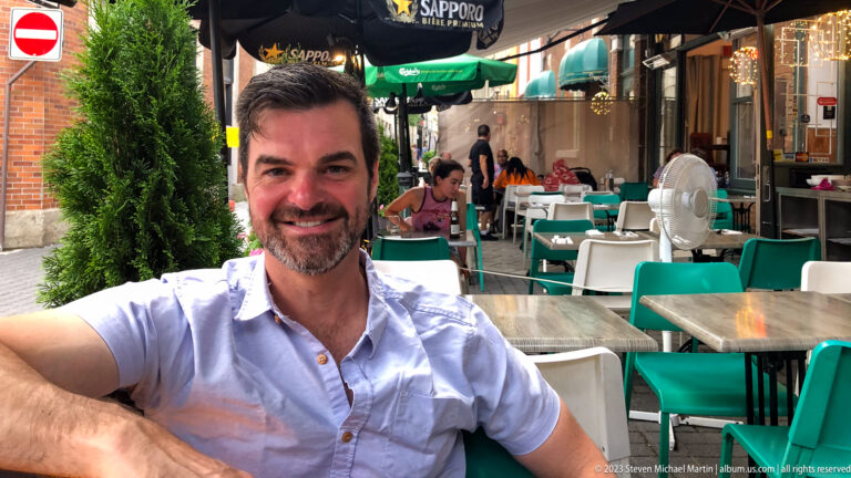 Smiling man in a light shirt seated at an outdoor restaurant terrace with turquoise chairs, tables, umbrellas, and diners behind.