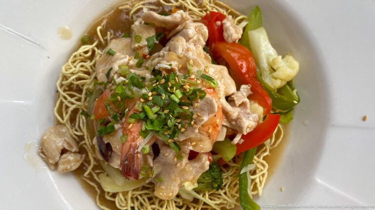 Crisp noodles topped with sliced chicken, red pepper, green vegetables, cauliflower, chopped herbs, and sesame seeds in a white bowl.