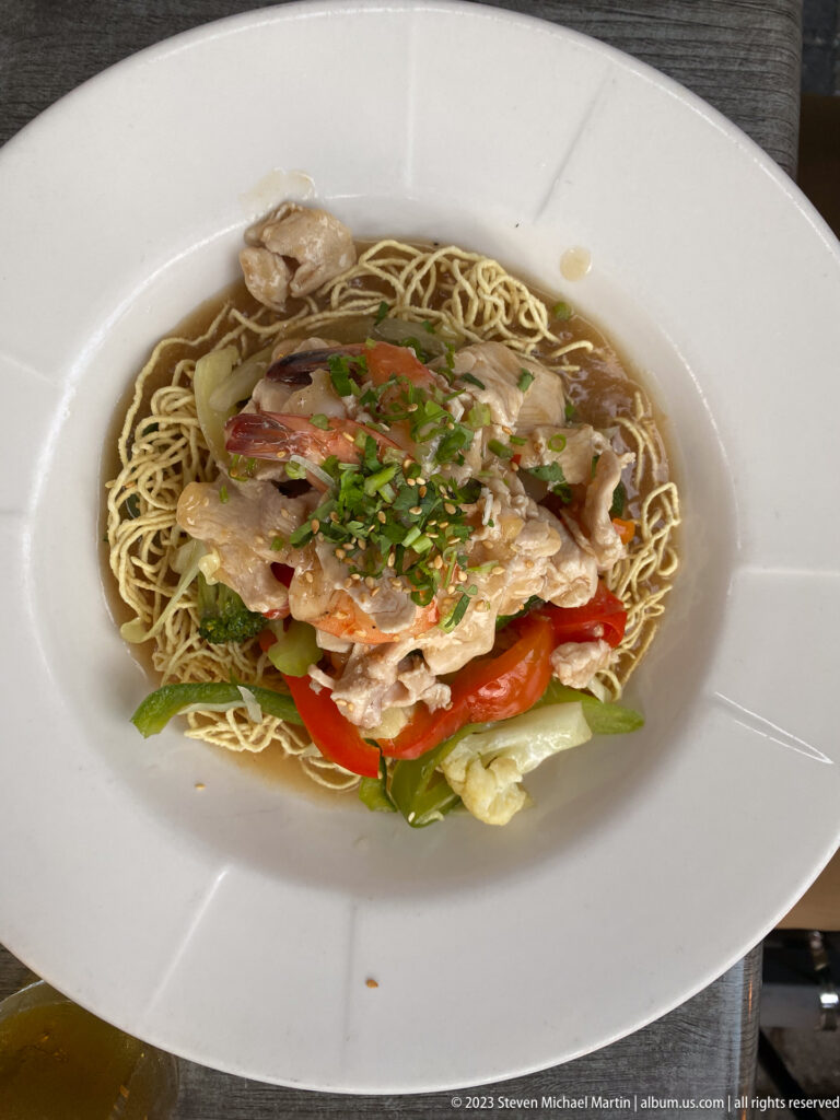 Wide white bowl holding crisp noodles topped with sliced chicken, red pepper, cabbage, chopped herbs, and sesame seeds.