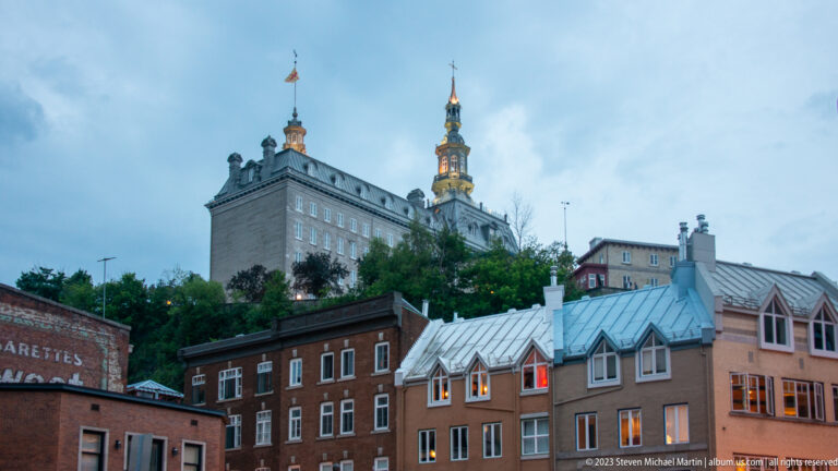 Large stone institutional building with clock and illuminated spired tower rising above brick buildings and metal roofs at dusk.