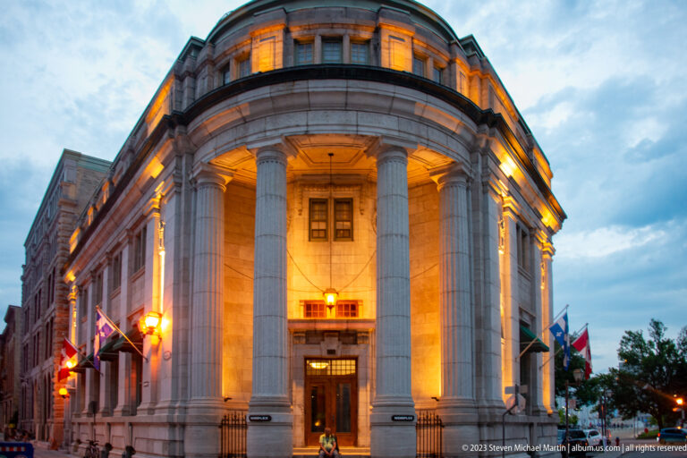 Curved pale stone civic building illuminated at dusk, with giant fluted columns, recessed entrance, upper windows, flags, and streetlights.