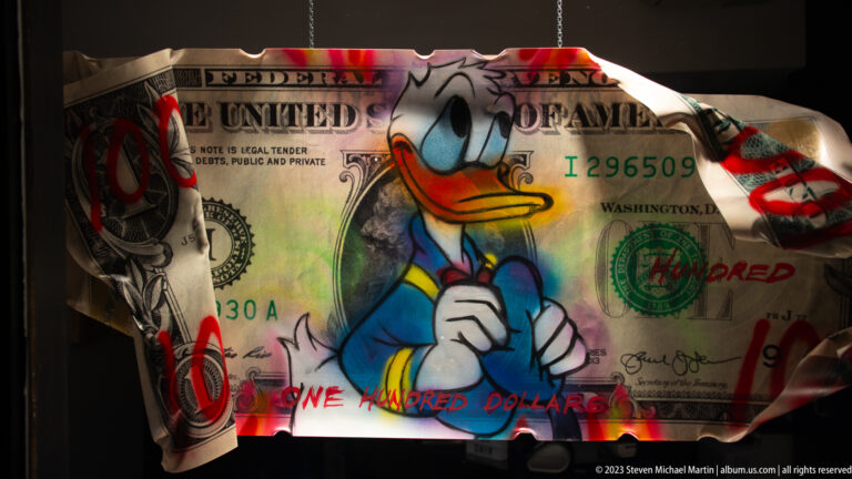 Close view of Donald Duck painted over a U.S. hundred-dollar bill with red circles, black marks, green paint, and handwritten words.