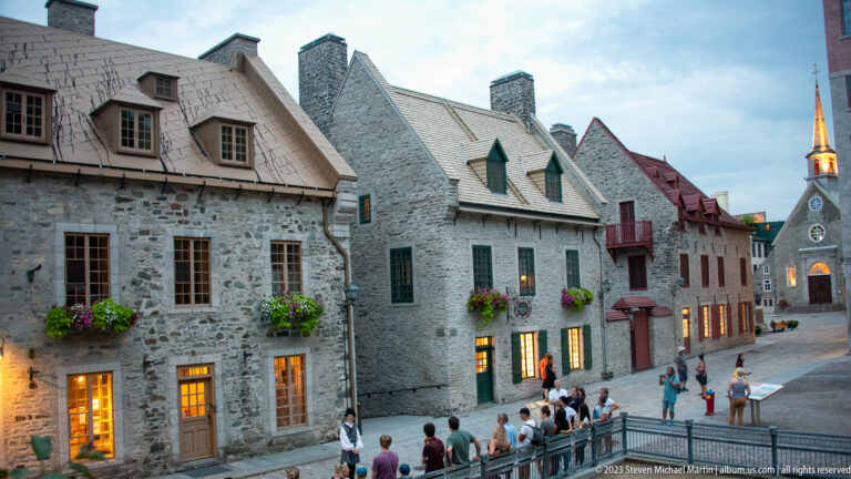 Historic stone houses with flower boxes line a sloping street leading toward the spired Notre-Dame-des-Victoires church.