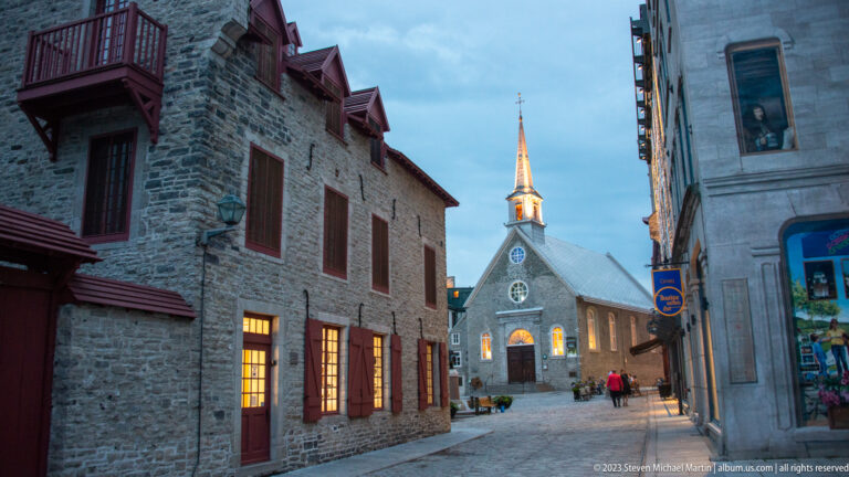 Notre-Dame-des-Victoires with illuminated windows and tall spire at the end of a narrow street between historic stone buildings.
