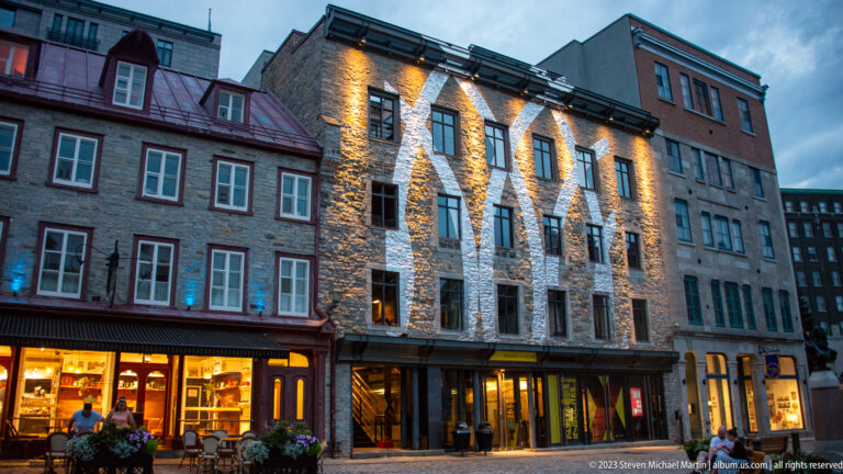 Illuminated multi-story stone building between historic commercial façades, with glowing shop windows and blue evening sky.