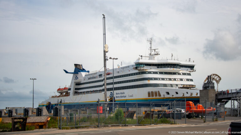 Large Marine Atlantic passenger-and-vehicle ferry berthed beside fenced terminal facilities under a cloudy sky.