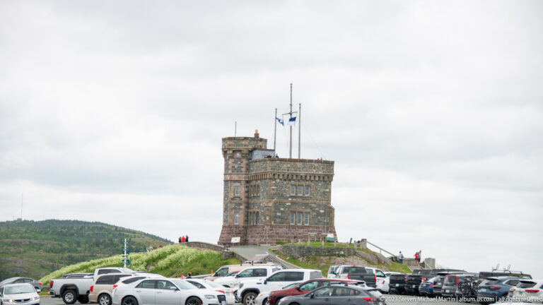 Cabot Tower above a busy Signal Hill parking area with cars, visitors, stone walls, and flagpoles.