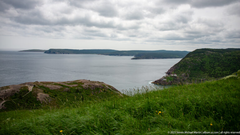 Green rocky headlands recede across gray-blue Atlantic water beneath heavy clouds, with tall grass in the foreground.