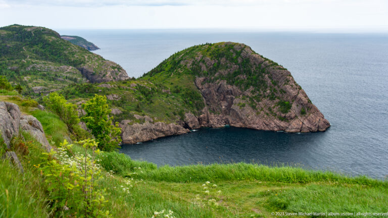 Rocky green headland enclosing a small dark-blue Atlantic cove below grassy slopes near Signal Hill.