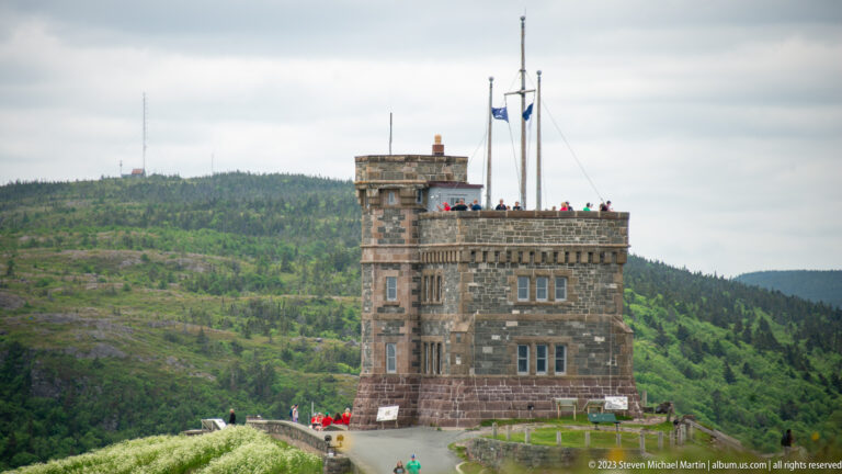 Telephoto view of Cabot Tower showing rough stone walls, crenellated roofline, windows, flagpoles, and visitors.