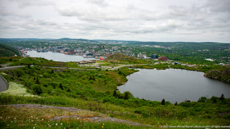 Dark pond and green Signal Hill slopes in the foreground with St. John’s harbor and city beyond.