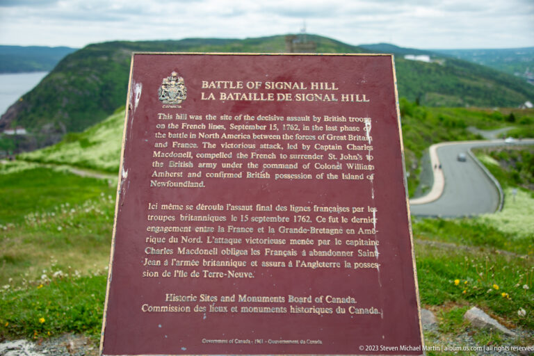 Bilingual Battle of Signal Hill commemorative plaque standing beside a winding road on the green hillside.