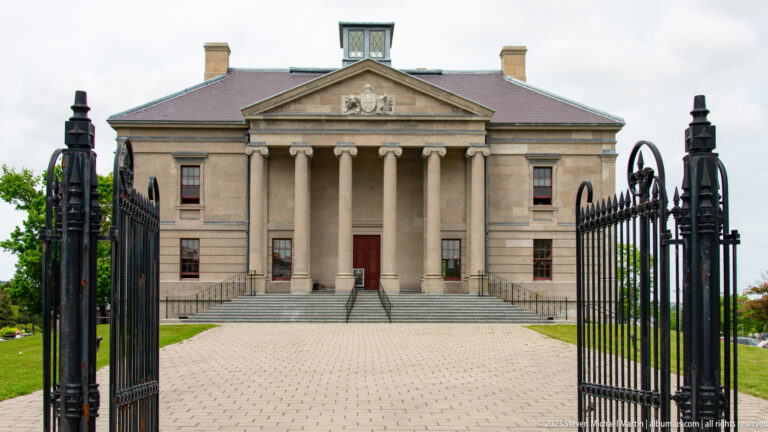 Colonial Building seen through open iron gates, with broad steps, six-column portico, and triangular pediment.