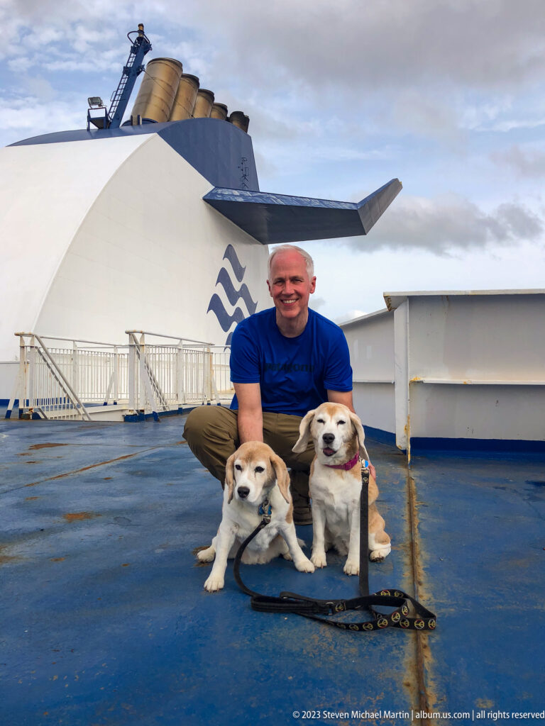 Man crouches behind two beagles on a blue ferry deck beneath the white Marine Atlantic funnel.