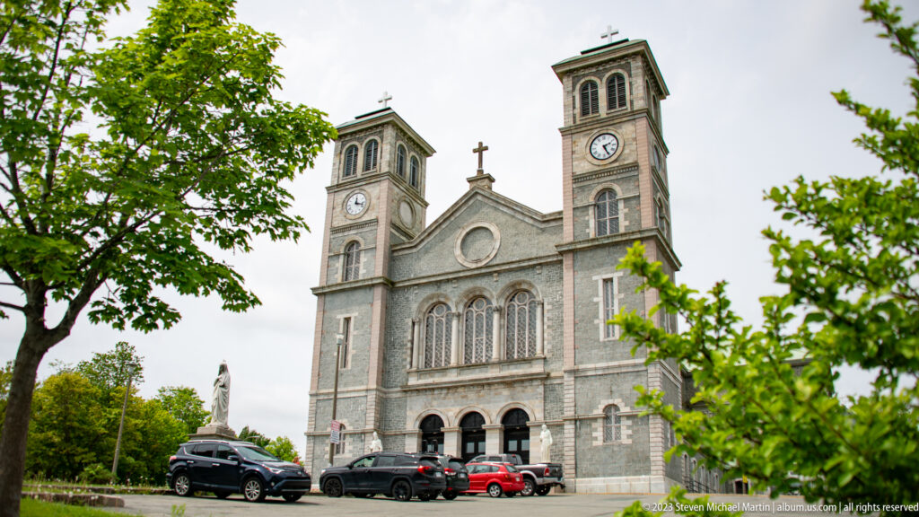 Basilica-Cathedral of St. John the Baptist with twin clock towers, central cross, arched windows, and gray stone façade.