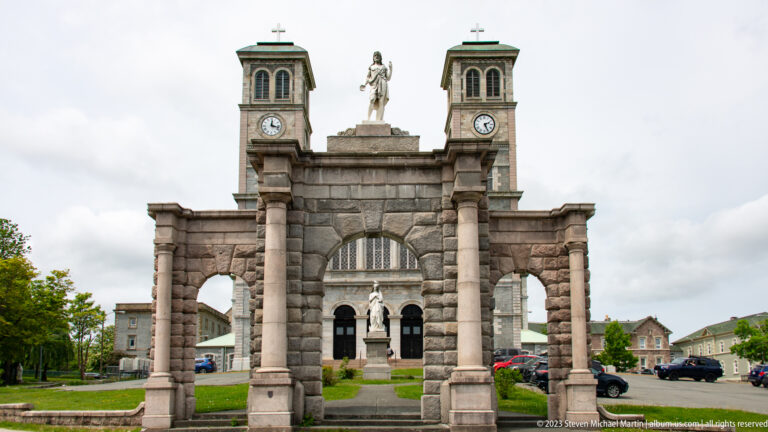 Three-arched stone gateway with a religious statue, framing the twin towers of the Basilica-Cathedral beyond.