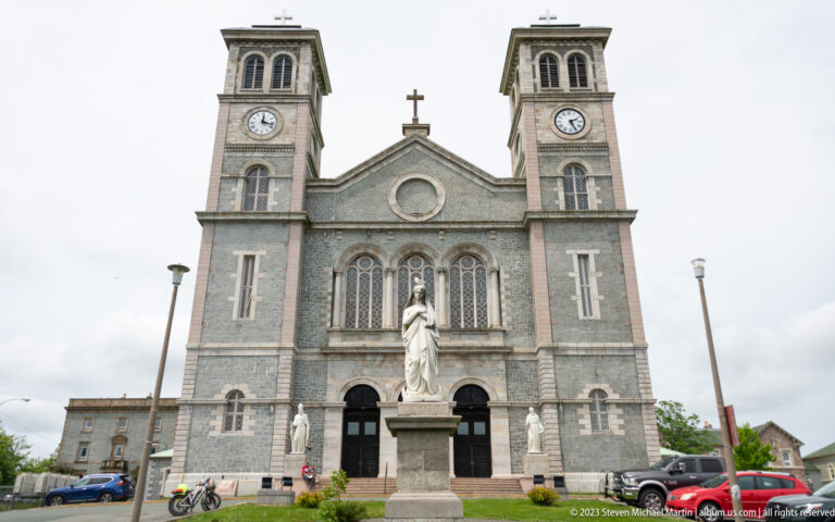 Frontal Basilica-Cathedral view with twin clock towers and a white Marian statue centered before the main entrance.