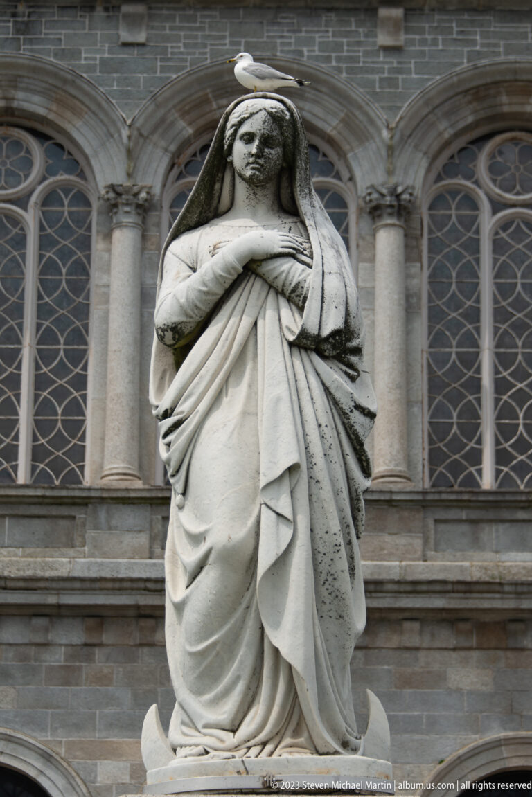Close view of a veiled stone Virgin Mary statue with crossed hands before tall arched cathedral windows.