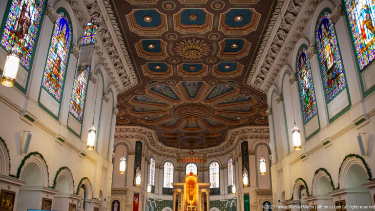 Wide Basilica-Cathedral interior with stained-glass windows, ornate painted ceiling, arches, hanging lights, and illuminated sanctuary.