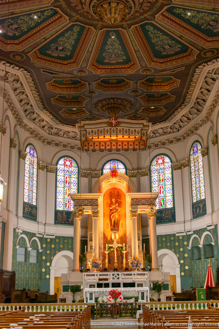 Symmetrical cathedral sanctuary with white altar, illuminated reredos, stained-glass windows, green patterned walls, and ornate ceiling.