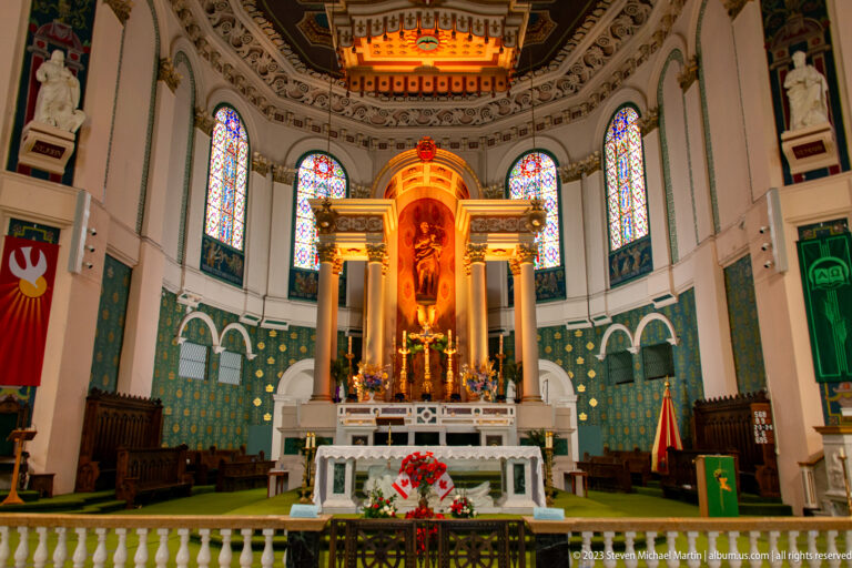 Cathedral sanctuary with white altar, glowing reredos, stained glass, colorful banners, flowers, and green patterned apse walls.