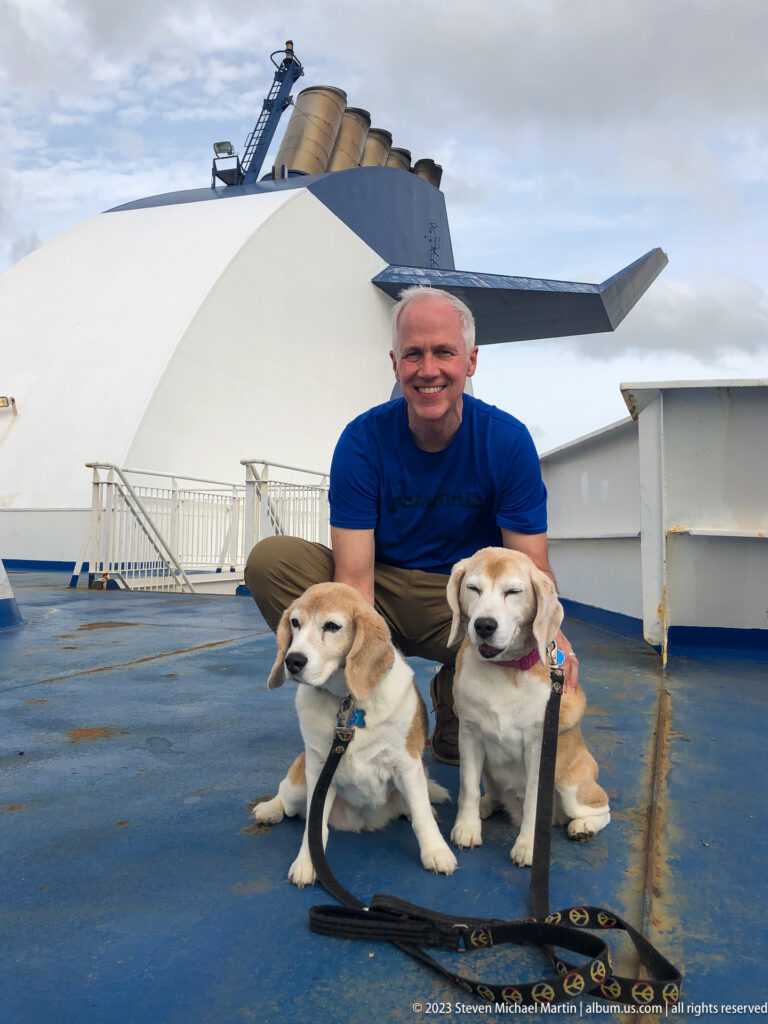 Smiling man poses behind two seated beagles on a blue ferry deck with the Marine Atlantic funnel in the background.