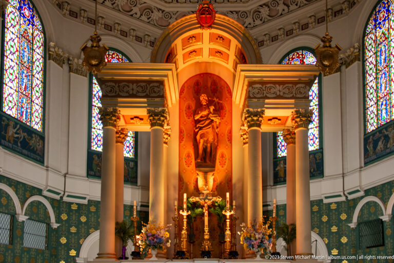 Illuminated religious statue in a sanctuary niche framed by columns, stained-glass windows, candles, and flowers.